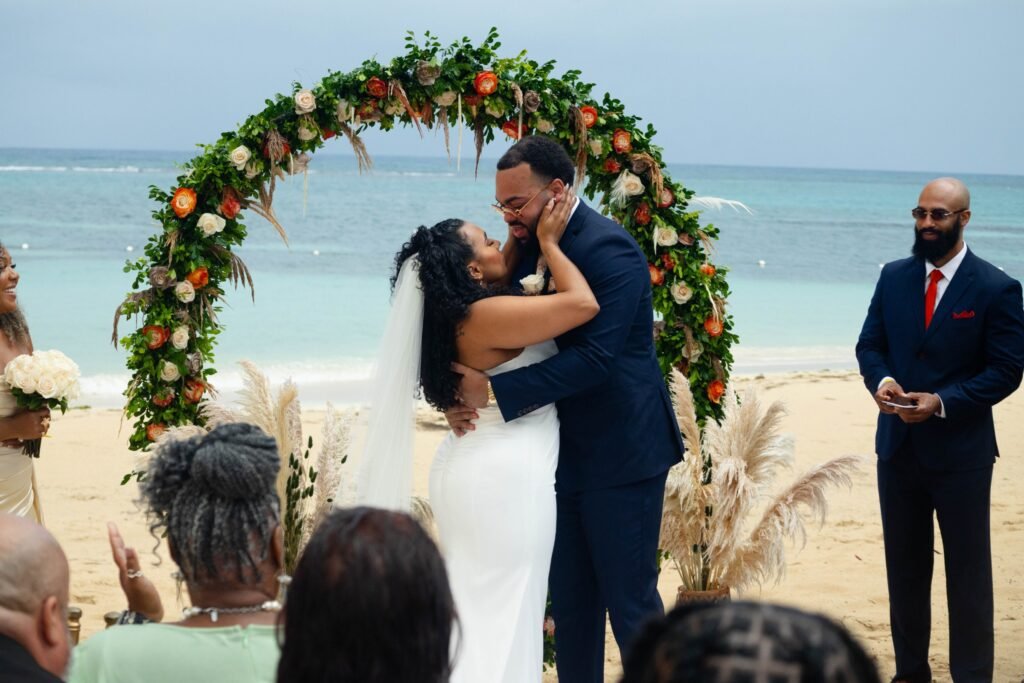 First kiss under the floral arch at a beach wedding ceremony — photographed by 121 Media Group
